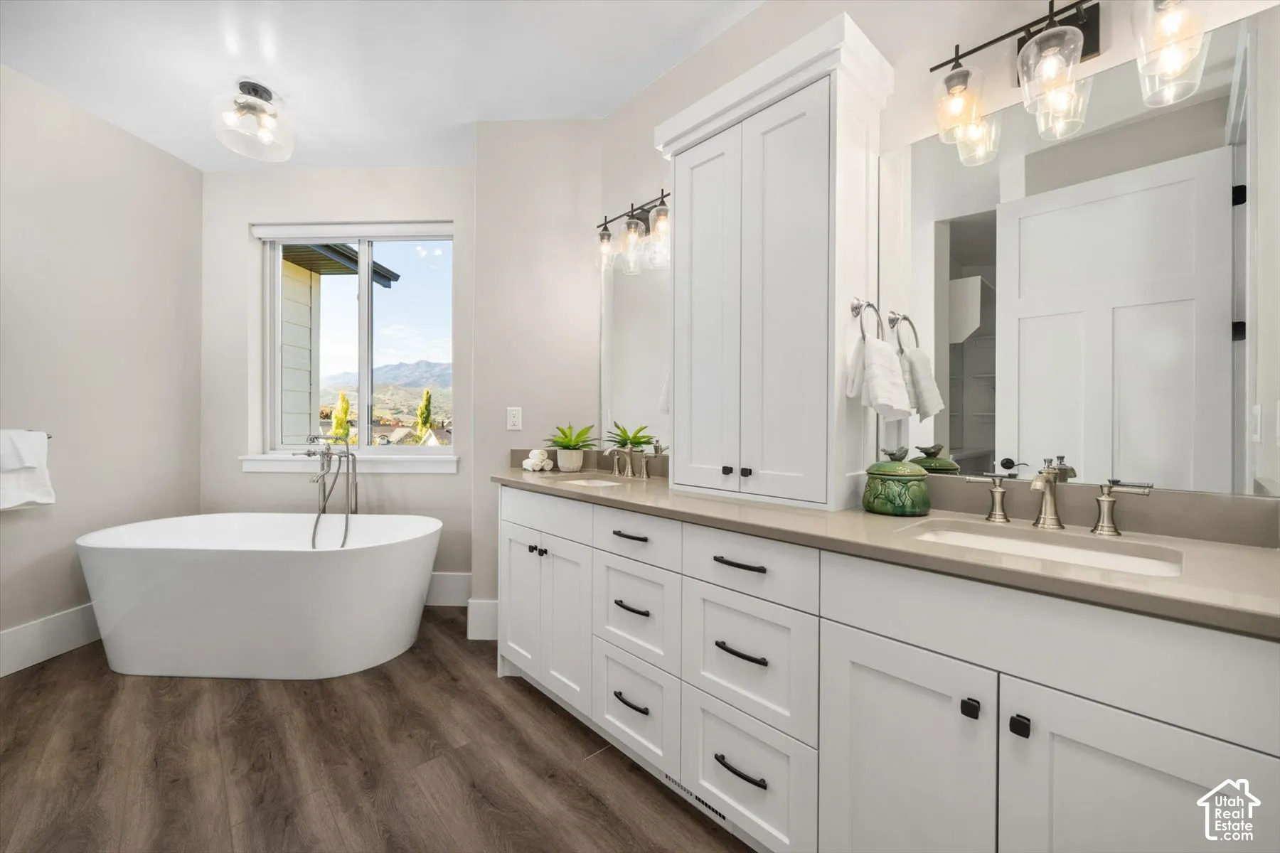 Freestanding soaking tub, dual vanity with undermount sinks, wood-finish flooring, custom white cabinetry, and a built-in linen tower
