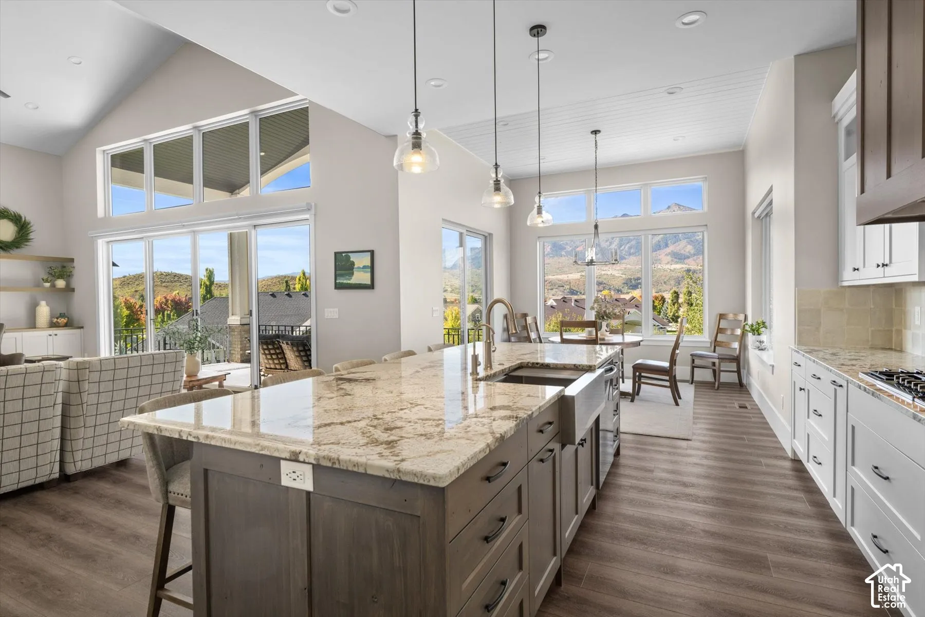 Open concept living area featuring vaulted ceilings, wood-finish flooring, a kitchen island with stone countertop and farmhouse sink, and a dining area with expansive windows