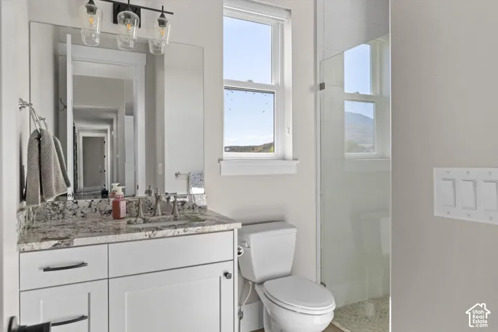 Bathroom featuring a vanity with a stone countertop, an undermount sink, a glass-enclosed shower, a toilet, and a window