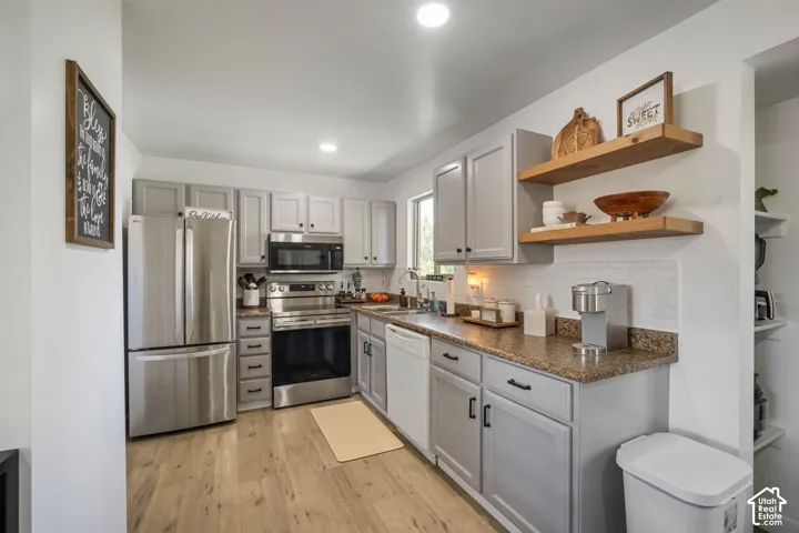 Light gray cabinetry with black hardware, stainless steel appliances, wood-finish flooring, white subway tile backsplash, and open wood shelving