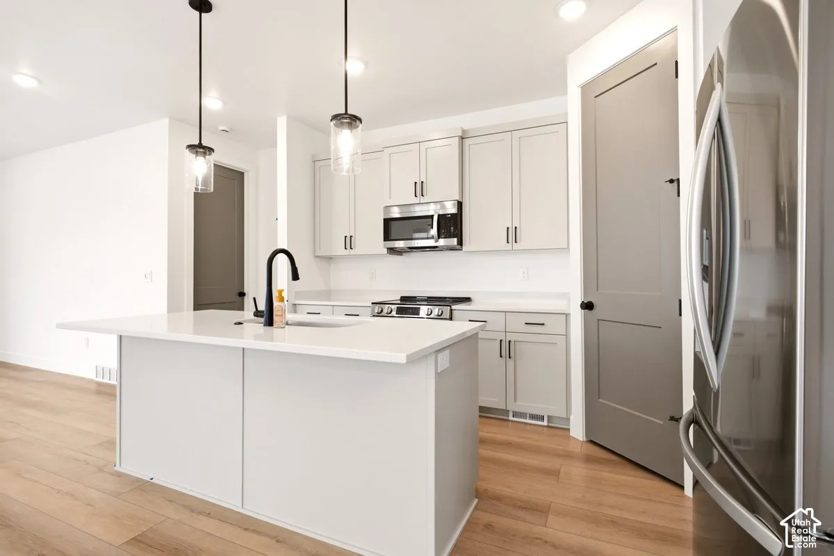 Kitchen featuring quartz countertops, an island with an undermount sink and matte black faucet, shaker-style cabinetry, stainless steel appliances, and wood-finish flooring