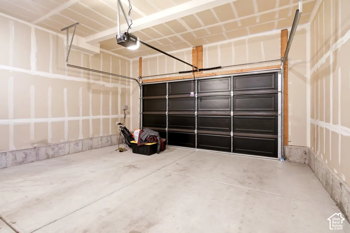 Concrete flooring, automatic garage door opener, dark-paneled garage door, exposed wood framing, unpainted drywall