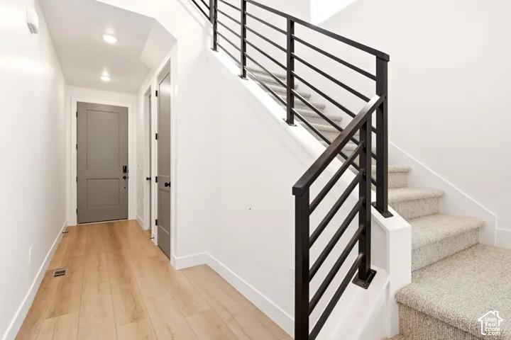 Entryway featuring light-toned wood-finish flooring, matte black hardware, stairwell with a metal horizontal-bar railing, and carpeted staircase
