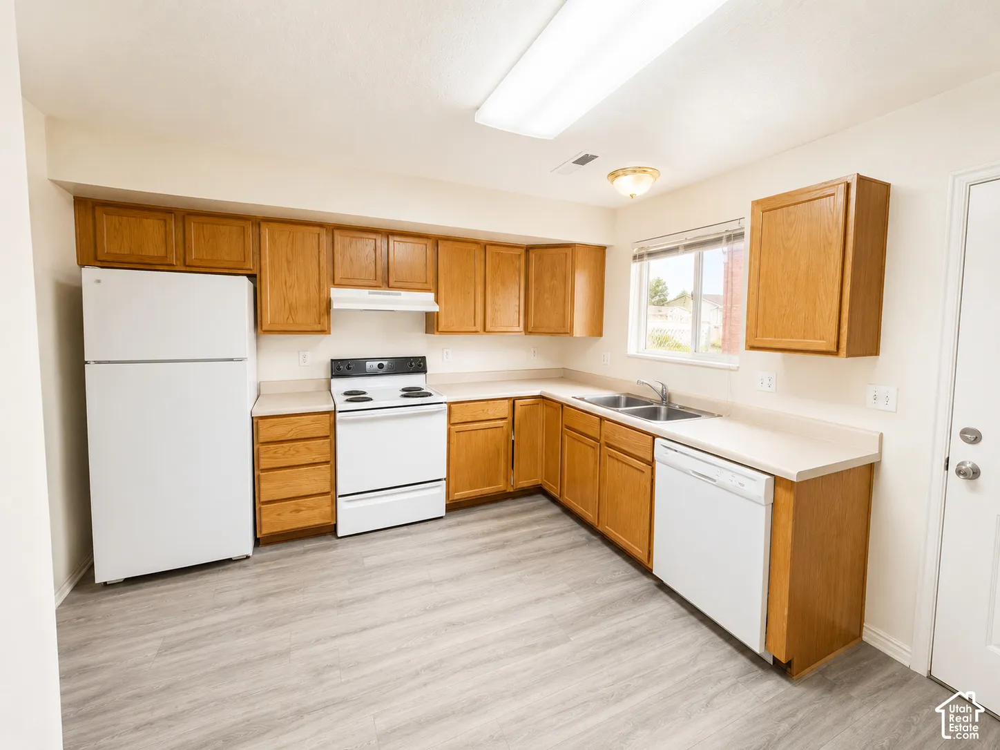 Spacious kitchen featuring wood-finish cabinetry, white appliances including a refrigerator and electric range, laminate countertops, a stainless steel sink with adjacent counter space, and wood-finish flooring throughout