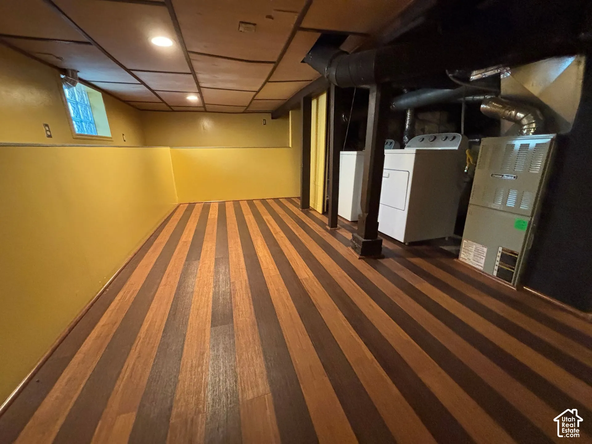Wood-finish flooring, drop ceiling with recessed lighting, and a utility area featuring a washer, dryer, and furnace