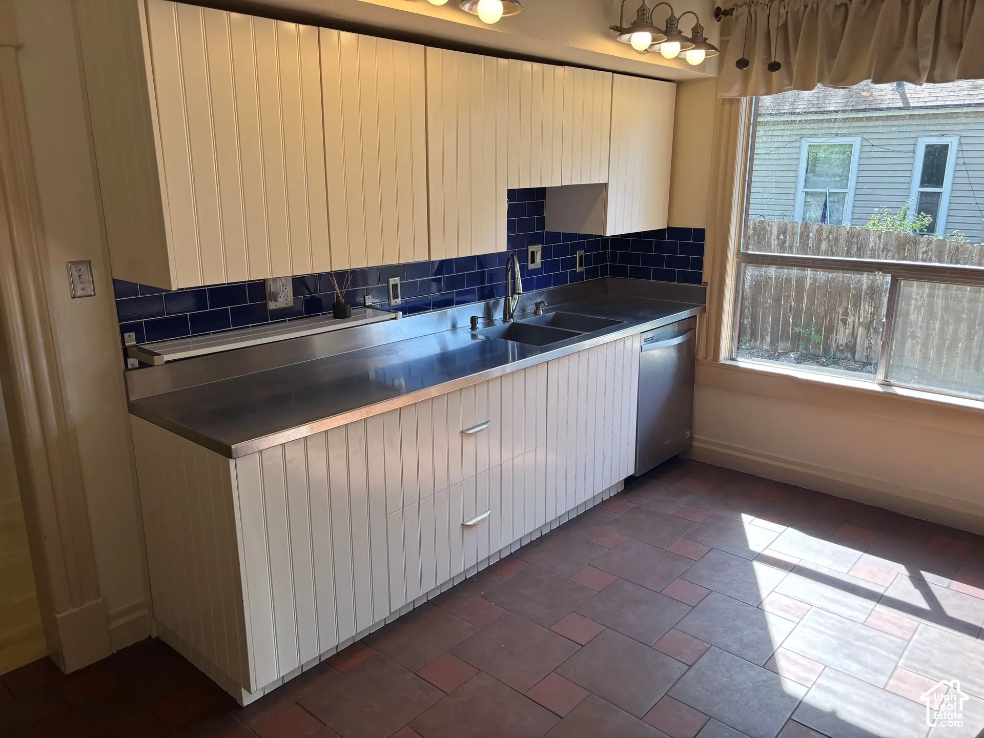 Kitchen featuring white beadboard cabinetry, stainless steel countertops with integrated double sink, blue subway tile backsplash, and a stainless steel dishwasher
