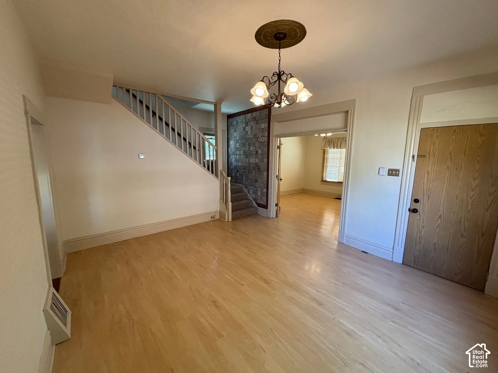 Light wood-finish flooring, staircase with painted balusters, decorative chandelier with ceiling medallion, wood-paneled door, and archway