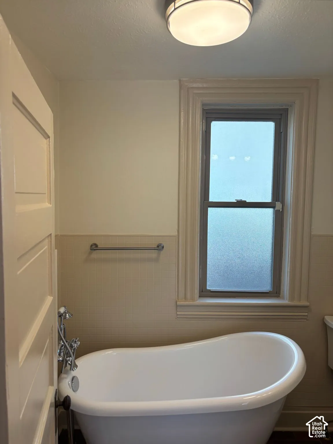Freestanding soaking tub with chrome fixtures, frosted window, and light-toned wall tile