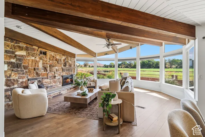 Vaulted wood-beamed ceiling, stone accent wall with fireplace, expansive window walls, wood-finish flooring, and a ceiling fan