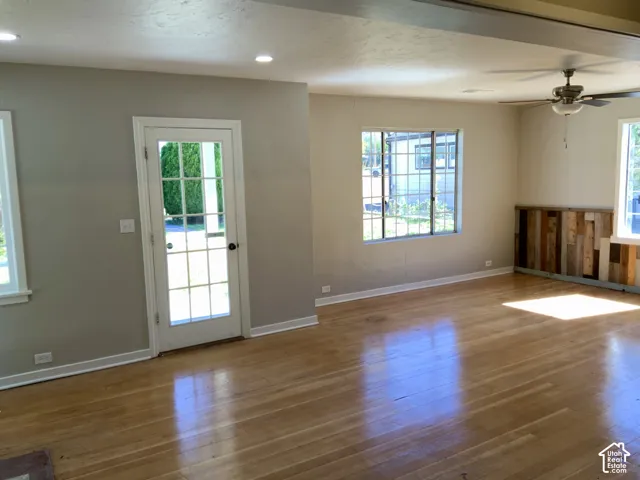 Spacious open-concept living area featuring gleaming wood-finish flooring, abundant natural light from multiple large windows and glass French door, neutral wall finishes, and recessed ceiling lighting