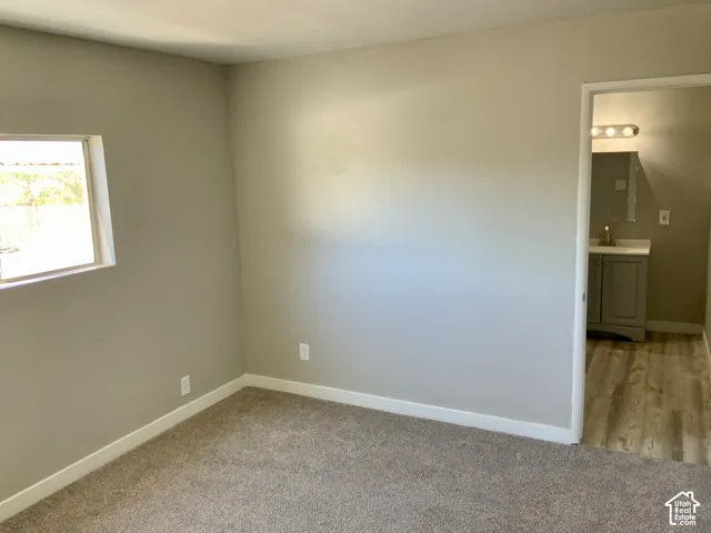 Carpeted room with a window, neutral wall tones, and an open doorway revealing a bathroom with a vanity, mirror, and wood-finish flooring
