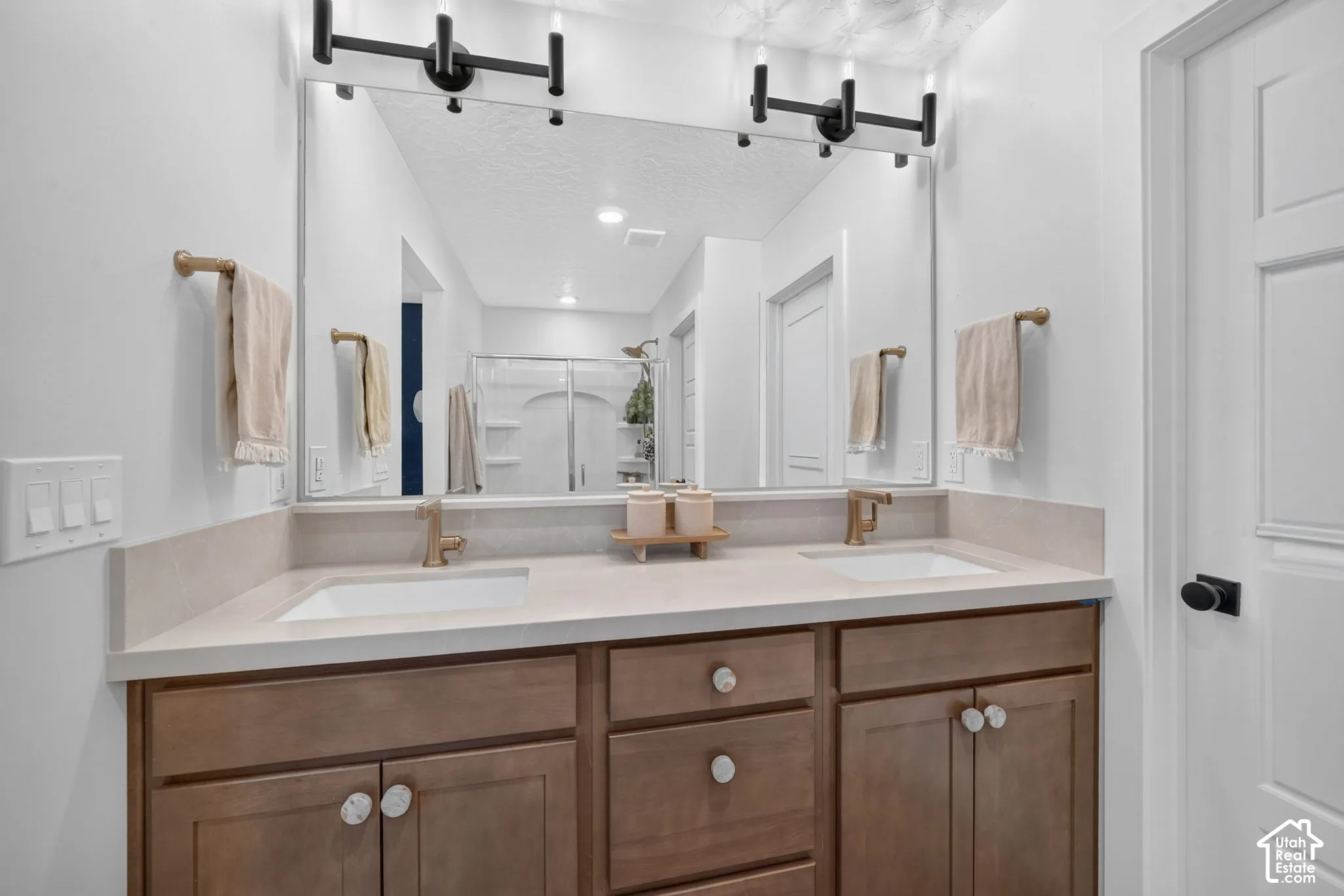 Dual vanity with integrated sinks, light-toned countertops, wood-finish cabinetry, and black-finish light fixtures