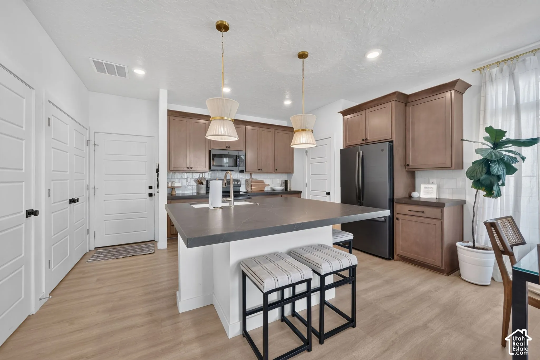 Kitchen featuring wood-finish cabinetry, black appliances, a central island with a sink, two pendant lights, and light wood-finish flooring