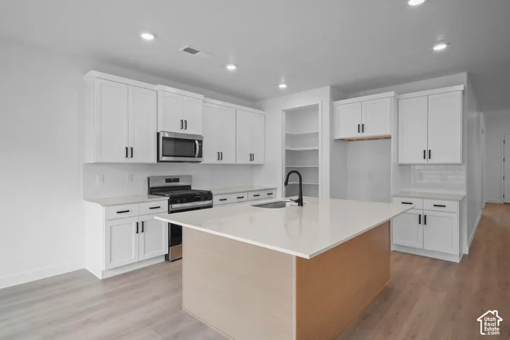 Modern kitchen featuring white shaker cabinetry, stainless steel appliances, a large center island with a sink and matte black faucet, and light wood-finish flooring