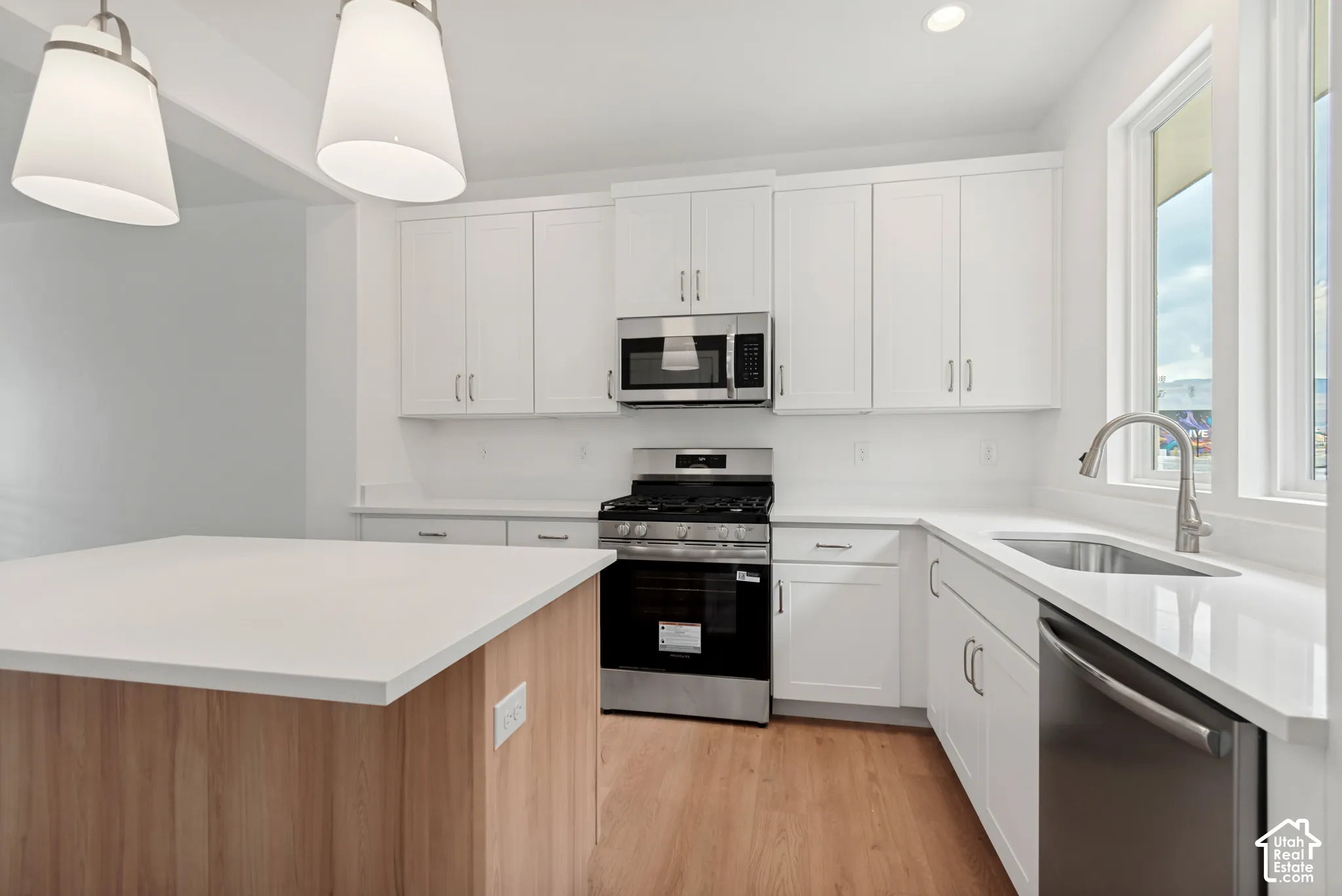 Modern kitchen featuring white shaker cabinetry, stainless steel appliances, light-toned wood-finish flooring, a kitchen island with a light countertop, and a stainless steel sink with a high-arc faucet