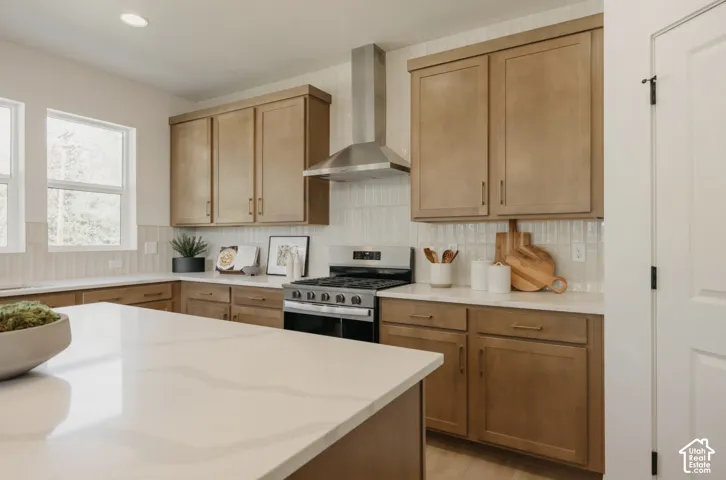 Kitchen featuring a gas range, stainless steel vent hood, light wood-finish cabinetry, light-toned countertops, and a white tiled backsplash