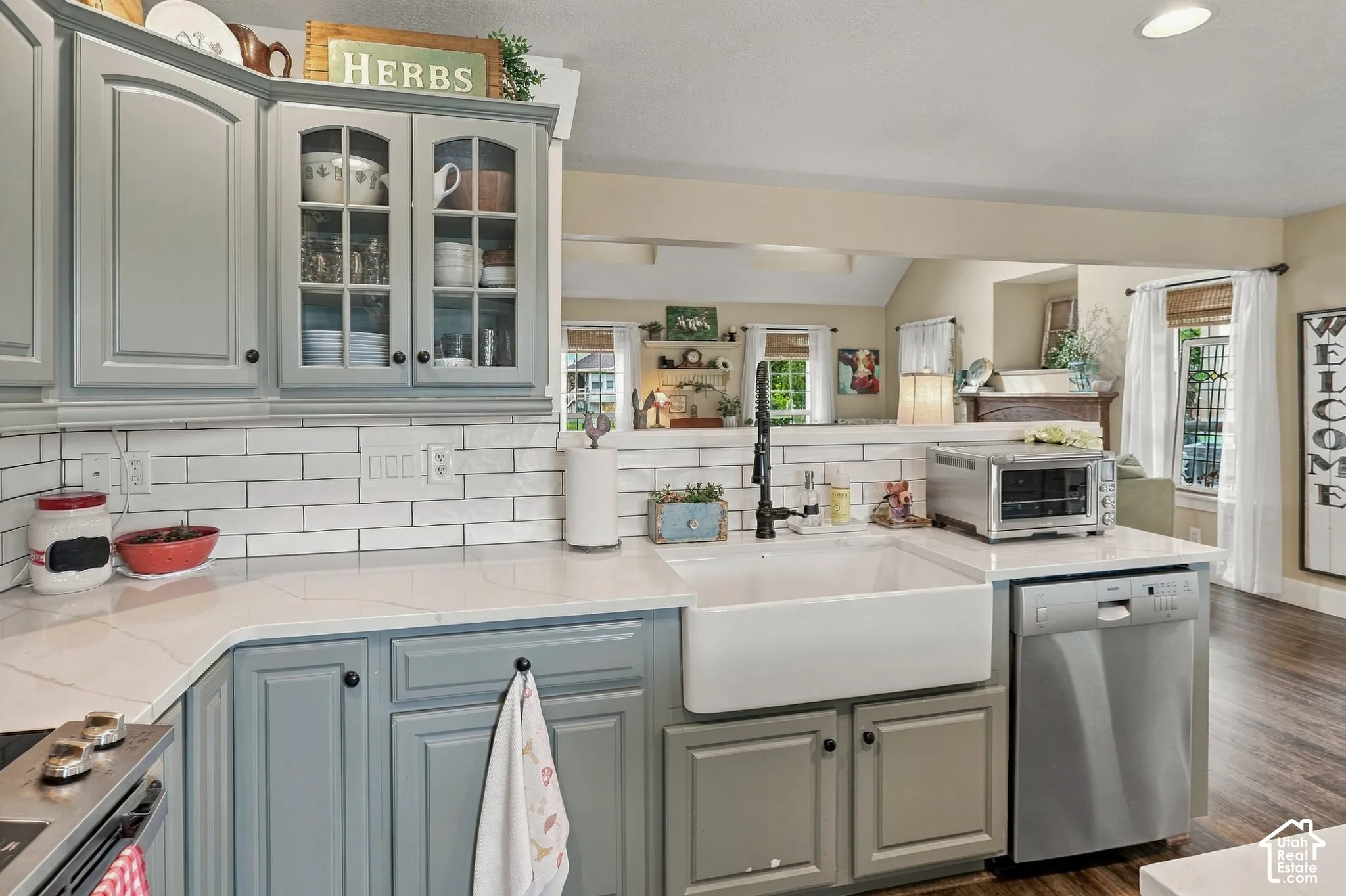 Kitchen featuring light gray cabinetry, white subway tile backsplash, white farmhouse sink, stainless steel dishwasher, and light-colored countertops