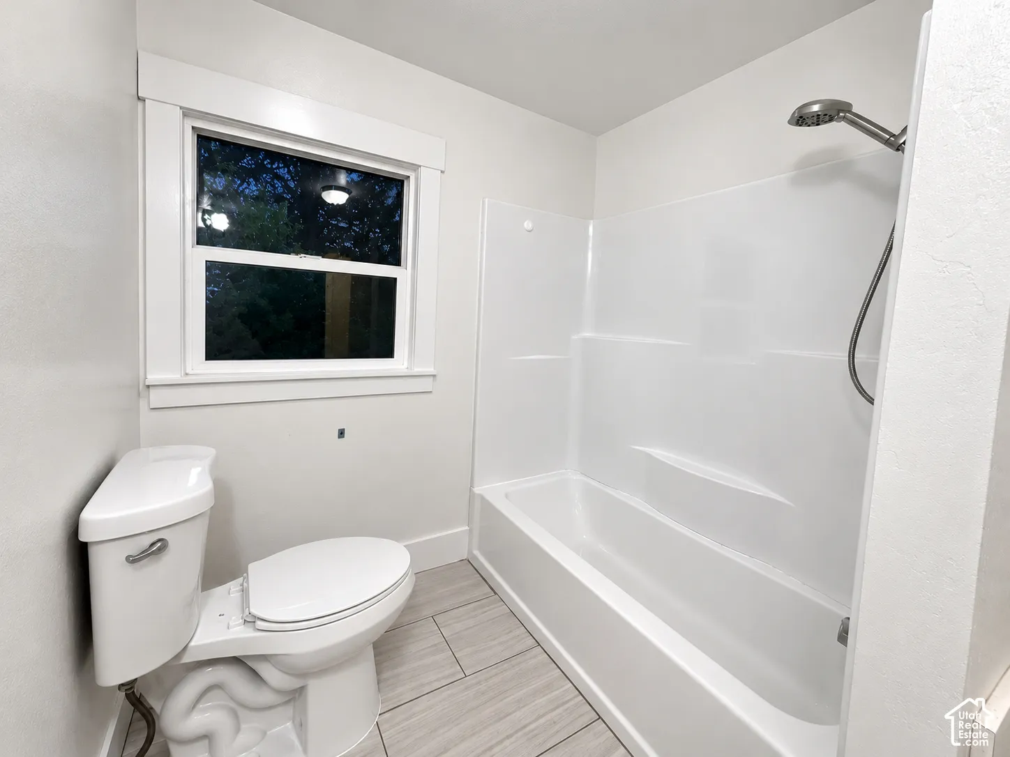 Bathroom featuring neutral tile flooring, a white acrylic tub-shower combination with a stainless steel handheld shower fixture, and a single-pane window with thick casing trim