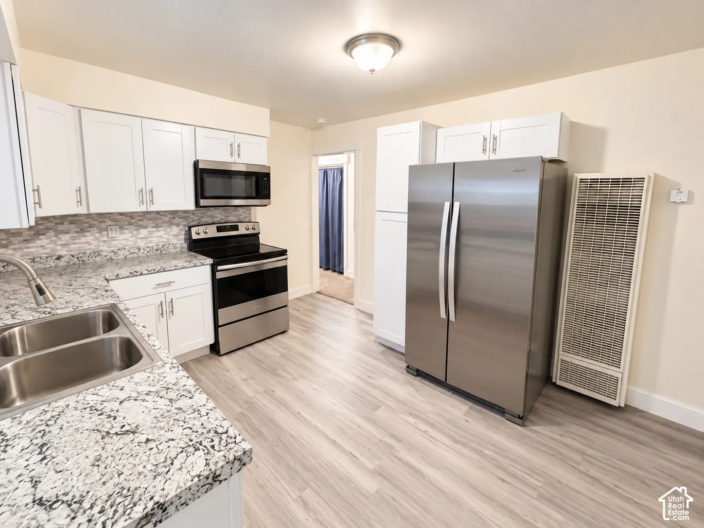 Kitchen featuring white shaker-style cabinetry, stone-finish countertops, stainless steel appliances, a multi-tone tile backsplash, and wood-finish flooring