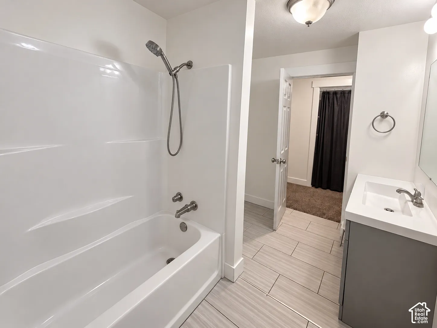 Bathroom featuring a white acrylic tub and shower surround, adjustable handheld shower fixture, gray vanity cabinet with integrated rectangular sink, and neutral-tone tile flooring