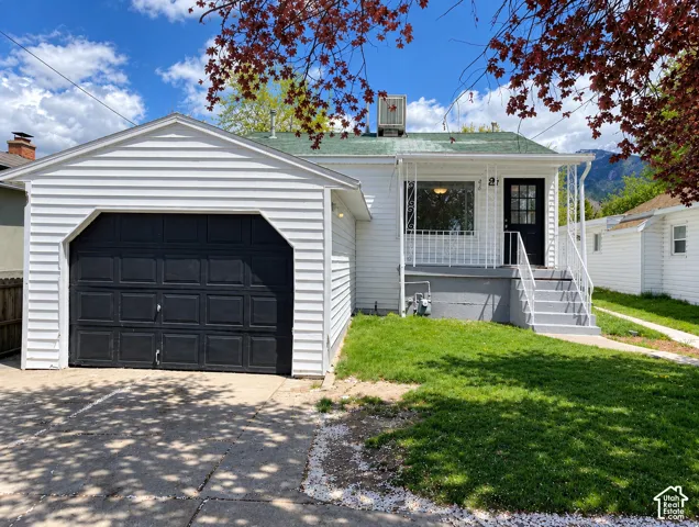 Exterior featuring white horizontal siding, attached single-car garage with black door, concrete driveway, front porch with metal railing, and mountain backdrop