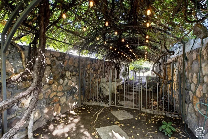 Overhead arbor with string lighting and substantial foliage, stone-clad walls, wrought-iron gate, and stone pavers