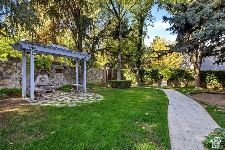 Manicured lawn with a stone pathway, a stone wall with an integrated fountain, a pergola, and mature trees