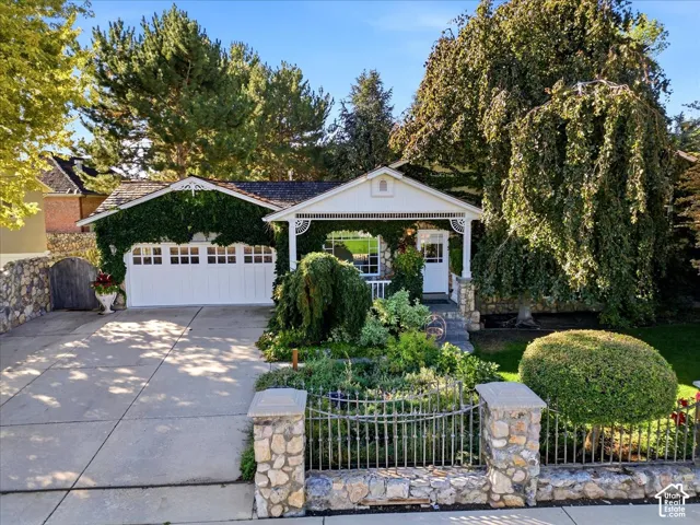 Ivy-covered facade with a prominent white garage door, stone wall accents, a covered front porch, and mature trees providing ample shade
