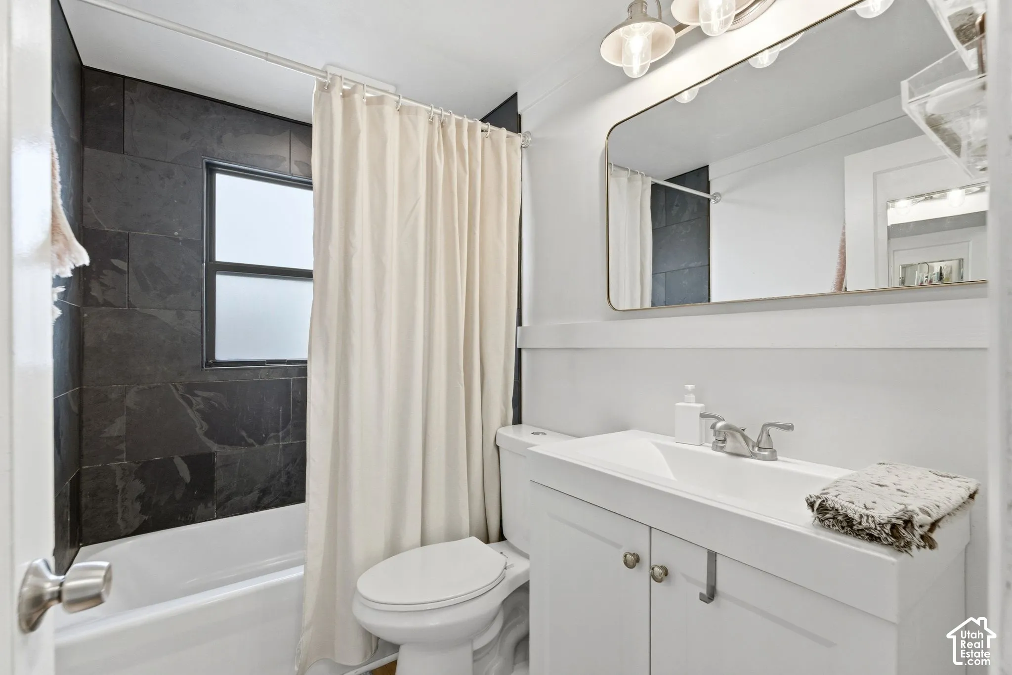 Tiled shower surround, frosted window, white bathtub, white vanity with integrated sink, and wall-mounted vanity lighting