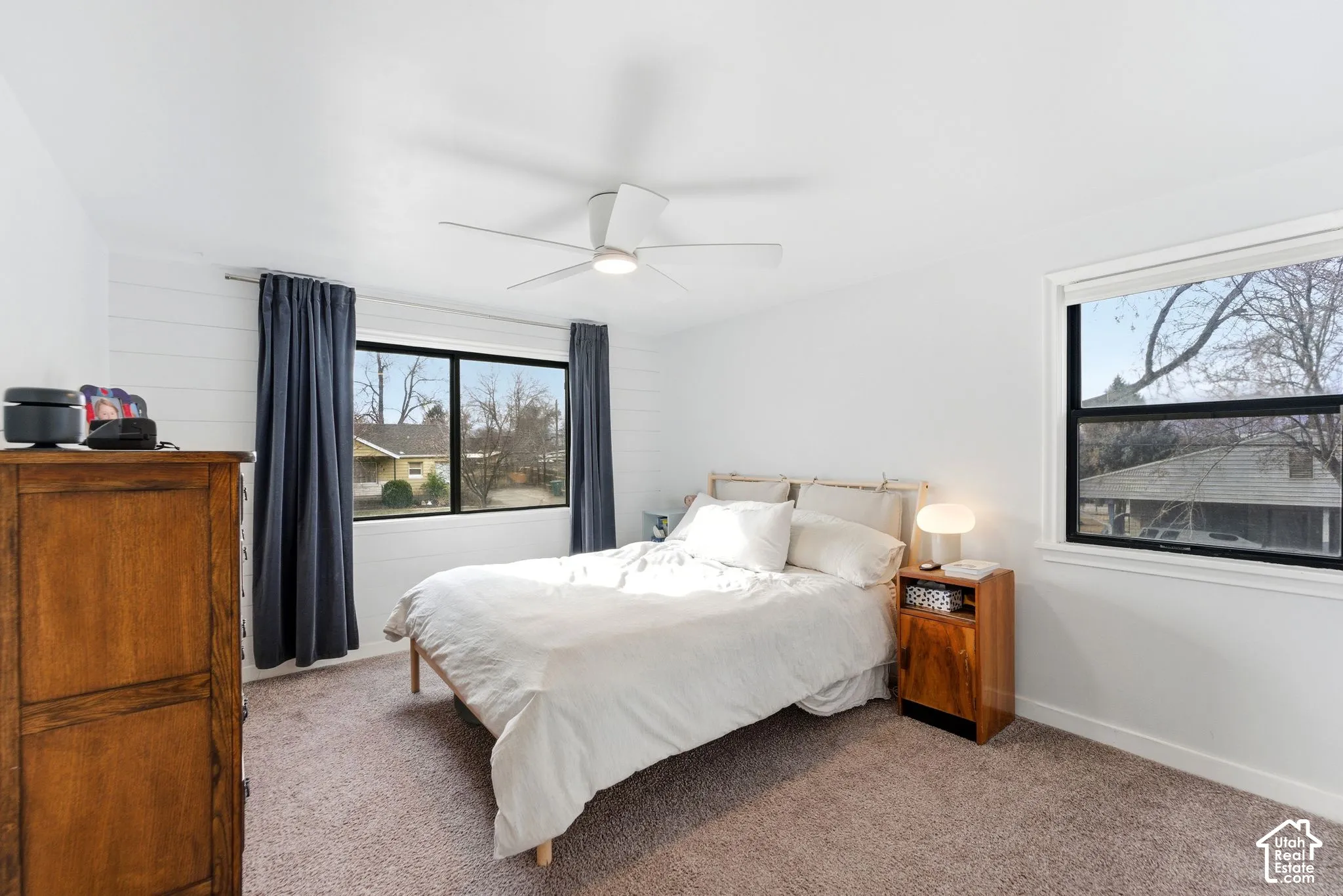 White-painted shiplap accent wall, two large windows with black framing, ceiling fan with integrated lighting, and carpeted flooring