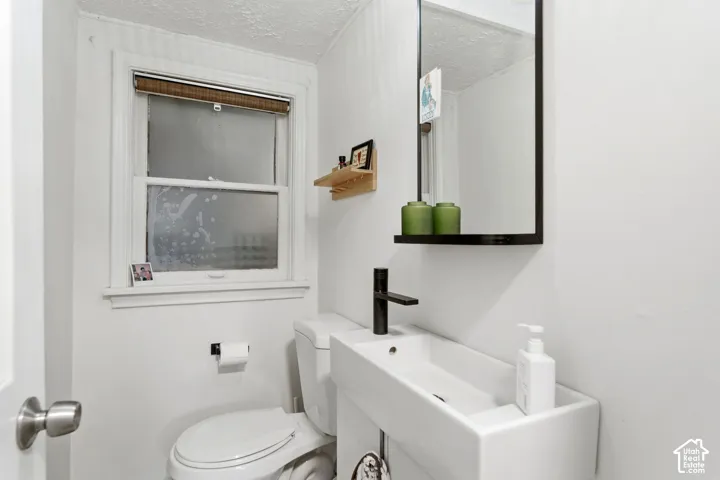White walls, rectangular window with a roller blind, white toilet, wall-mounted rectangular basin, and a black framed mirror with an integrated shelf