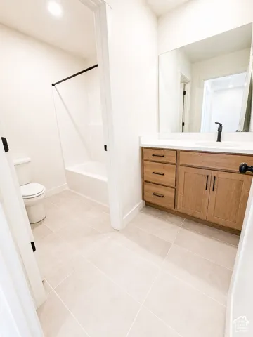 Light-toned tile flooring, single vanity with light wood-finish cabinetry, white countertop, integrated sink with matte black faucet, and white tub/shower combination with black hardware