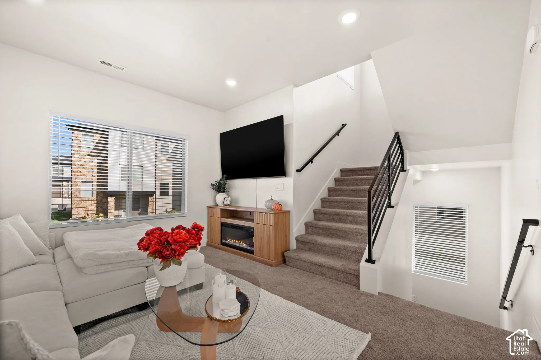 Recessed lighting, wood-finish media console, integrated electric fireplace, carpeted stairs with metal railings, and a large window with blinds