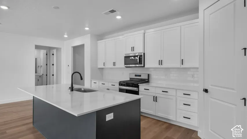 Modern kitchen featuring an island with a matte black faucet and under-mount sink, white shaker cabinetry with dark hardware, stainless steel appliances, white subway tile backsplash, and wood-finish flooring