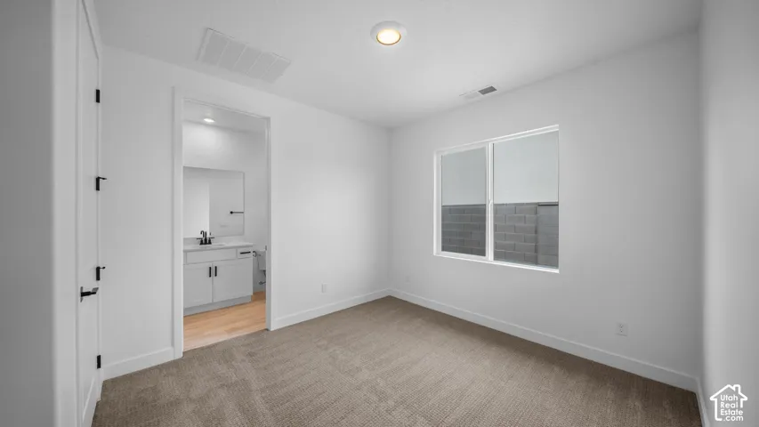 Carpeted room with white walls, a single window, and a doorway revealing a bathroom with a white vanity, a mirror, and wood-finish flooring