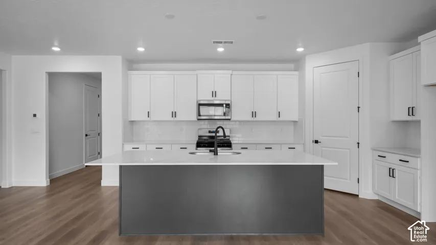 Modern kitchen featuring a dark gray island with an integrated sink, white shaker cabinetry, stainless steel appliances, and wood-finish flooring