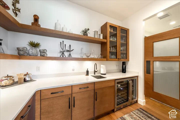Wood-finish floating shelves, white countertops, built-in beverage refrigerator, wood-finish cabinetry, and a stainless steel sink with a black faucet