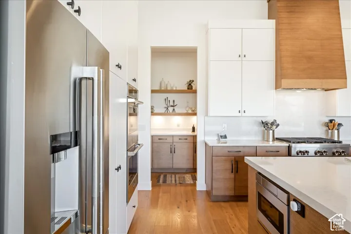 Stainless steel refrigerator, integrated double ovens, hardwood flooring, white countertops, and a wood-finish range hood