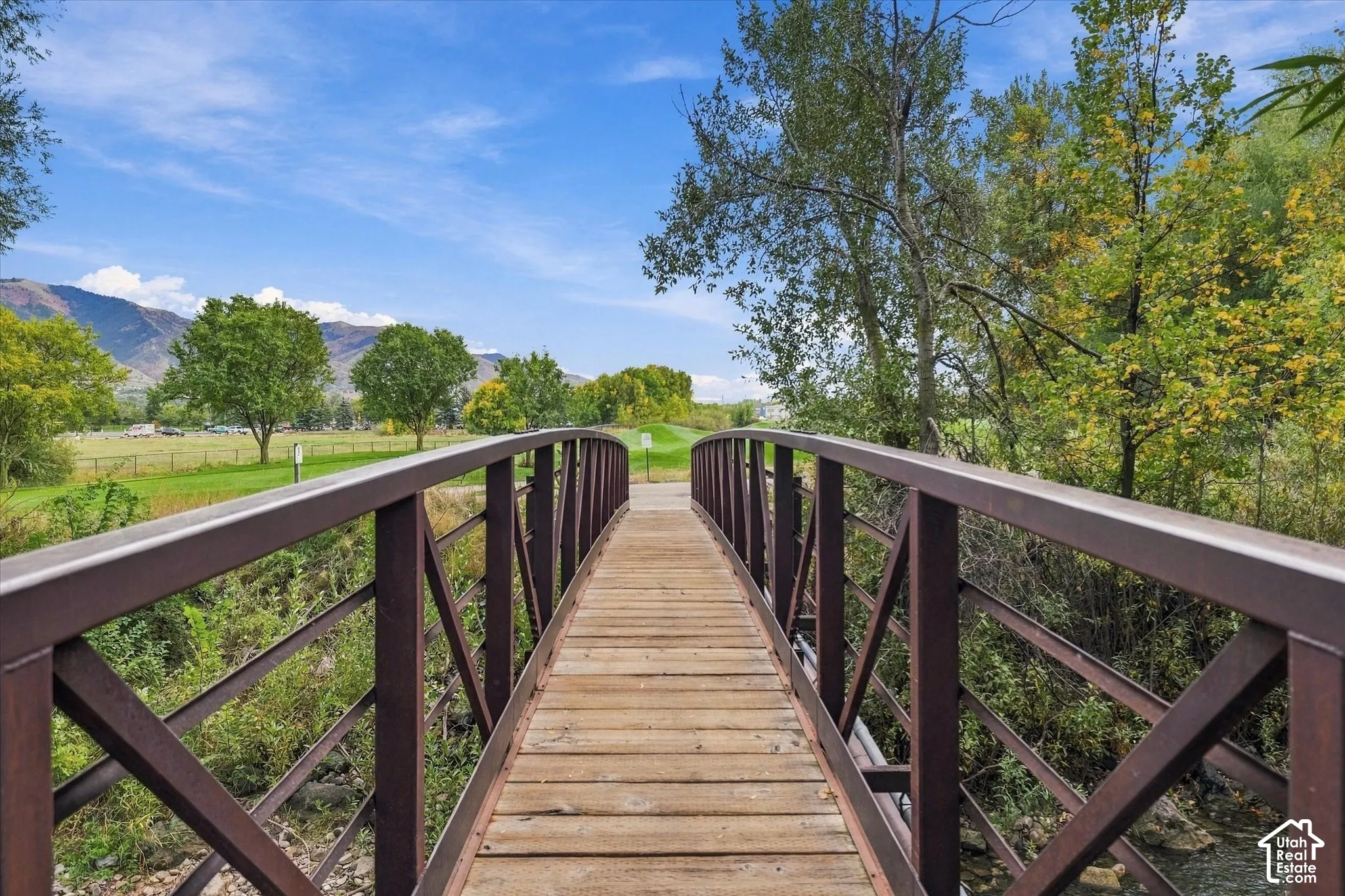 Wooden bridge with metal railings over a small waterway, surrounded by lush trees and a grassy expanse