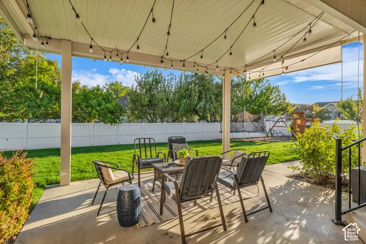 Covered patio with string lighting, concrete flooring, and a white privacy fence enclosing a grass lawn