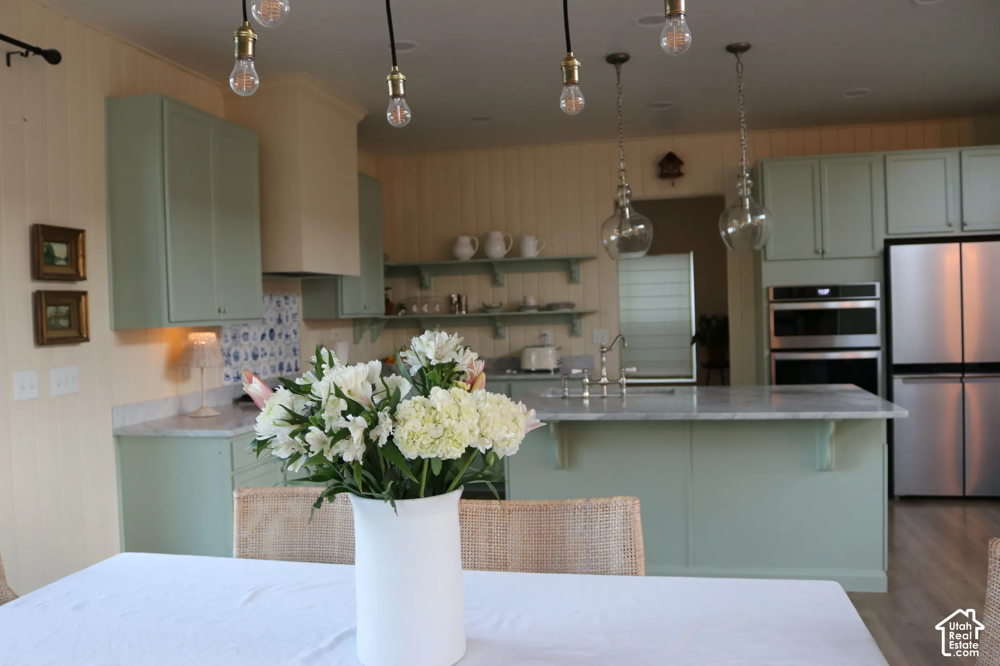Kitchen featuring light green cabinetry, a center island with a sink, pendant lighting, stainless steel appliances, and wood-finish flooring