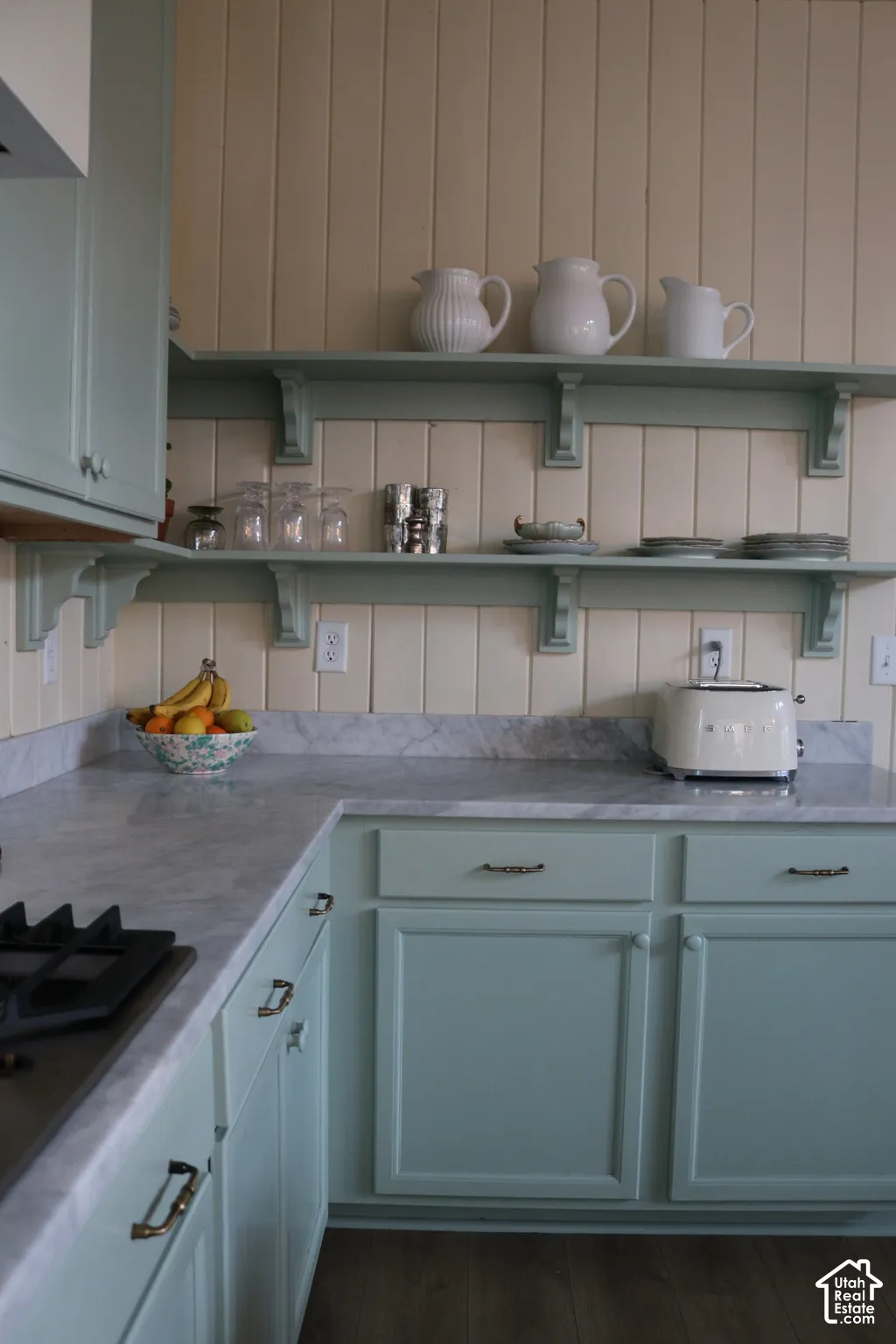Mint green cabinetry with decorative pulls, integrated gas cooktop, light marble-finish countertops, and open shelving