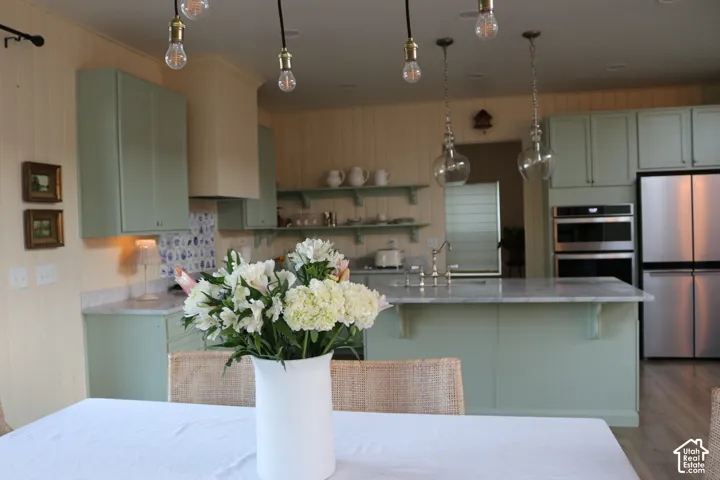 Kitchen featuring light green cabinetry, a center island with a sink, pendant lighting, stainless steel appliances, and wood-finish flooring