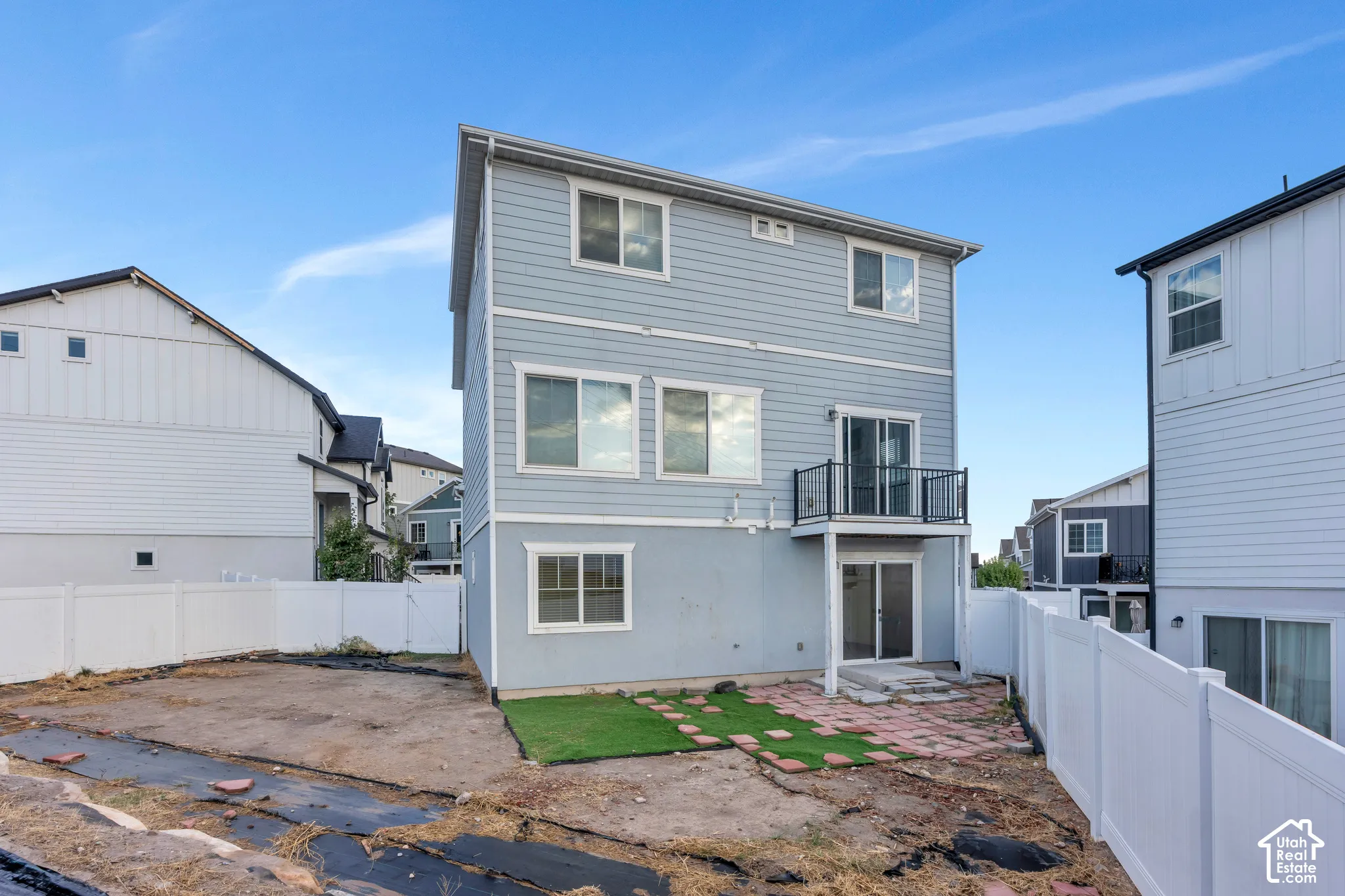 Two-story residential structure with light blue siding, a ground-level patio, second-story balcony with glass railing, and white perimeter fencing