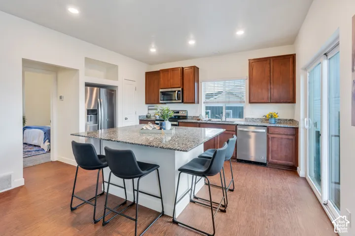 Kitchen featuring a central island with a speckled countertop, rich wood-tone cabinetry, stainless steel appliances, and wood-finish flooring