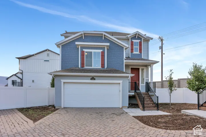 Two-story property featuring a front-facing garage, brick paver driveway, blue siding, red entry door, and exterior stairs with black railings