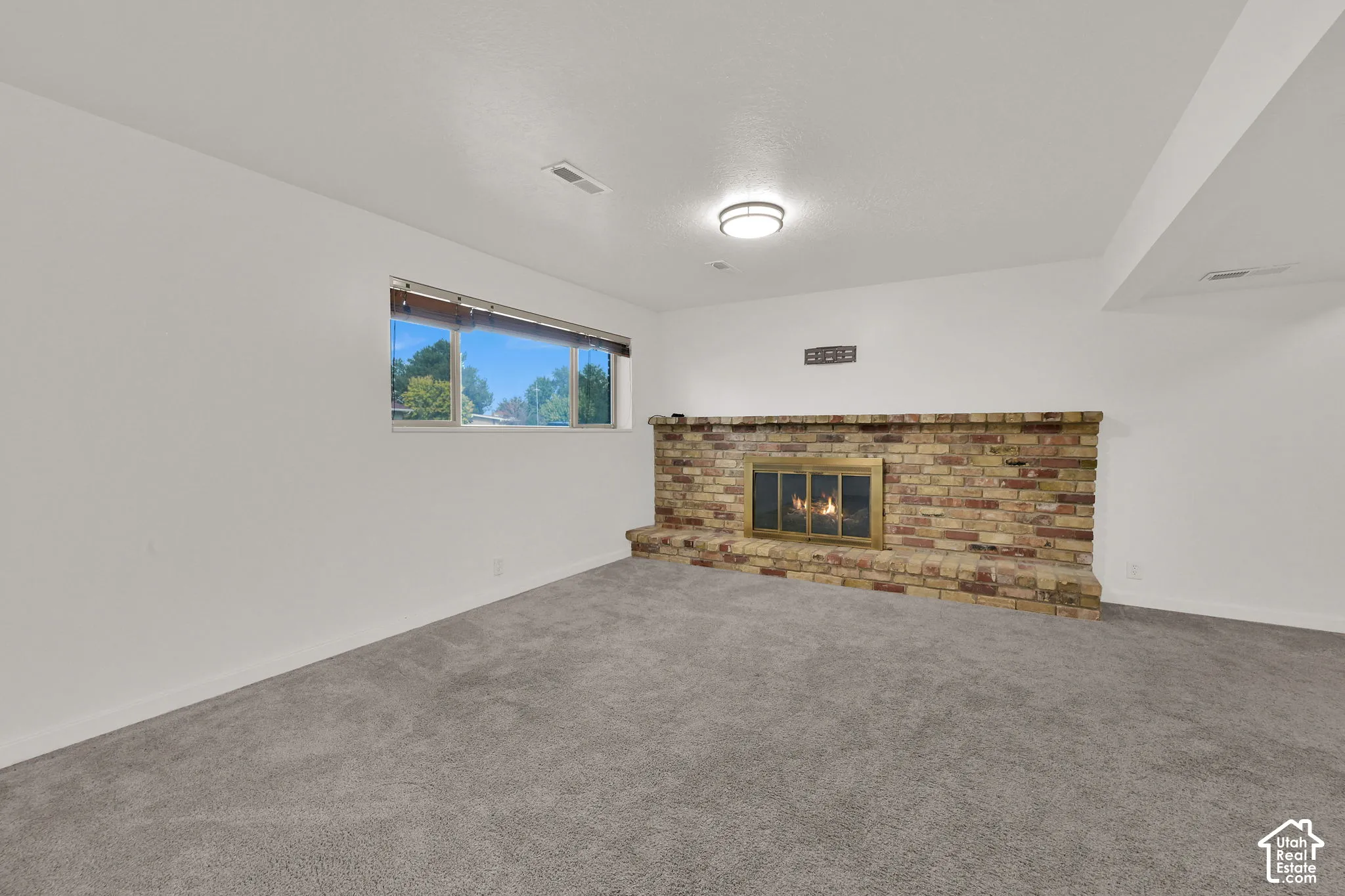 Brick fireplace with a brass firebox, flush-mount ceiling light, white walls, and gray carpeting
