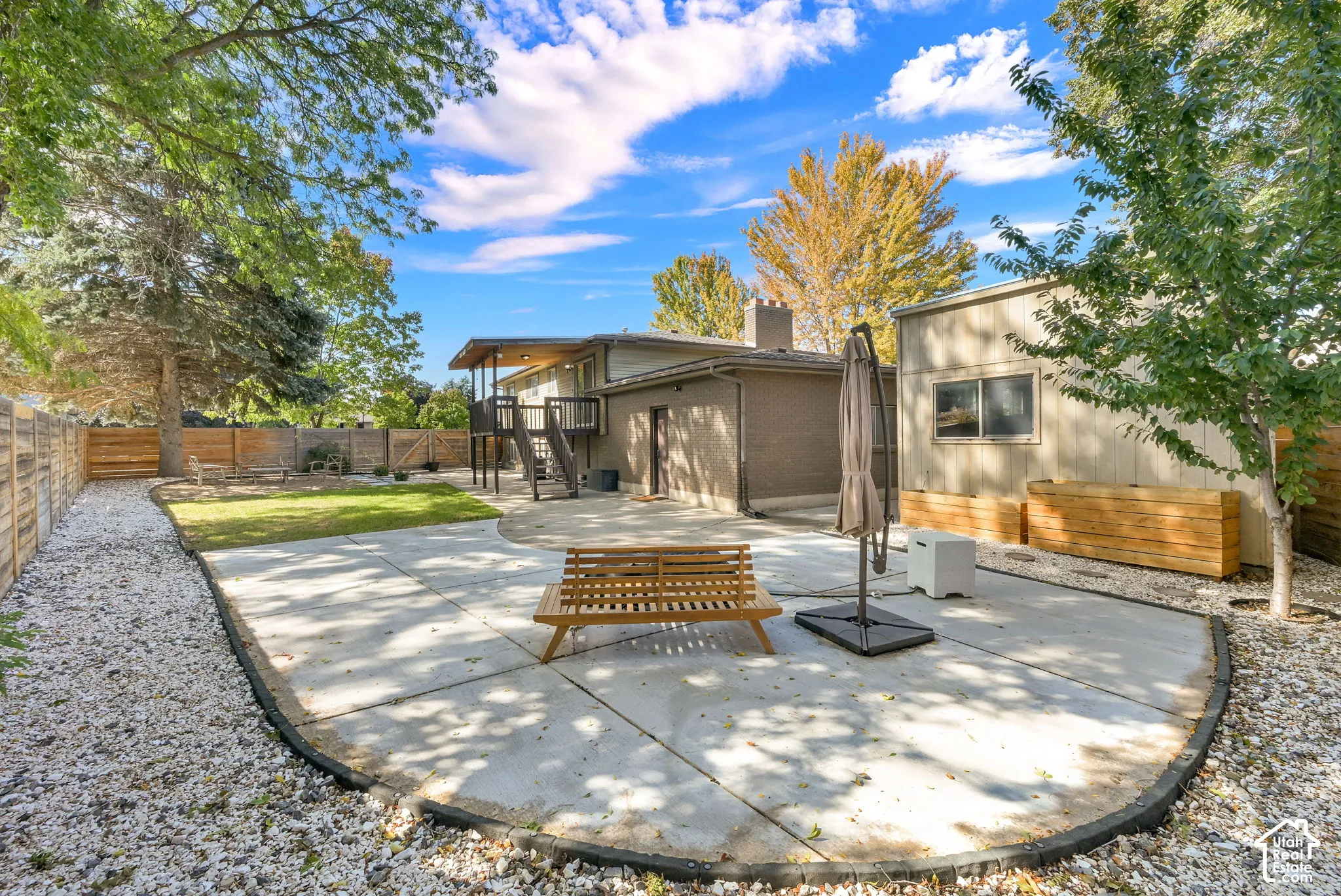 Expansive backyard featuring a large concrete patio, grassy area, gravel landscaping, a raised wooden deck, and mature trees