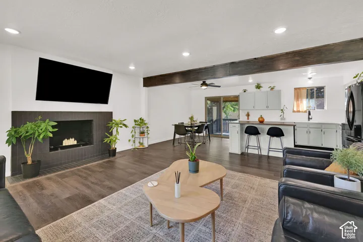 Spacious living area featuring wood-finish flooring, a fireplace with dark tile surround, exposed ceiling beams, and recessed lighting