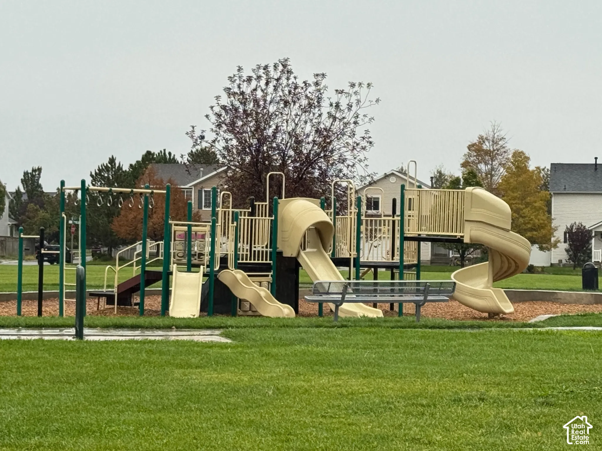Community playground featuring multiple slides, climbing structures, and swings, surrounded by a manicured lawn and mature trees